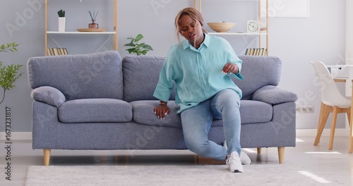 Woman faints while sitting on sofa at home. Young African American girl, sitting on couch in living room, suddenly feels unwell and falls down unconscious on floor