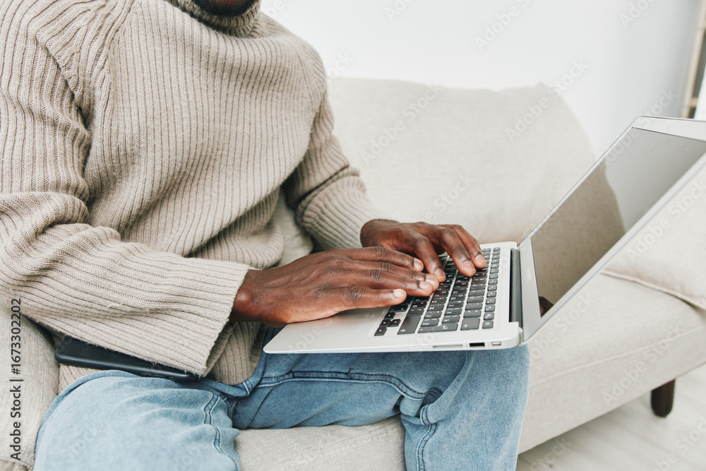 Naklejka premium A young Black man working on a laptop while seated on a cozy couch, wearing a beige sweater and casual jeans, depicting a relaxed and productive home environment
