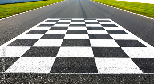 Close up view of a black and white checkered finish line on a paved asphalt race track with green grass on either side