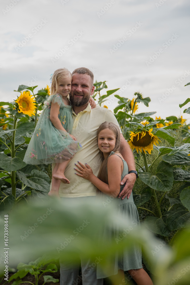 Fototapeta premium A father and two daughters dressed in yellow and green clothes are standing in a bright sunflower field, enjoying a sunny day in the fresh air, creating joyful memories of the beauty of nature.
