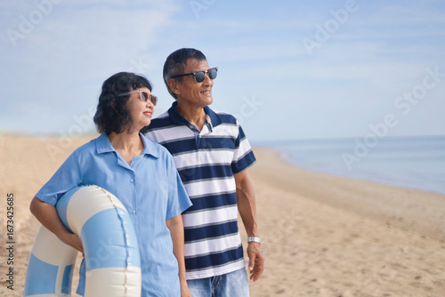 Cheerful senior couple traveler wearing a casual shirt and holding swimming ring in front of the morning Beach with copy space.