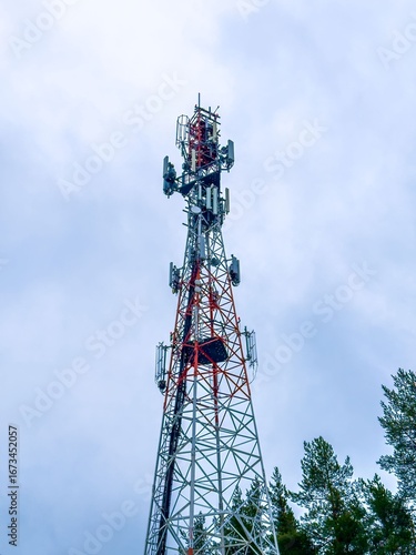 A dramatic, low-angle shot of a tall telecommunication tower with red and white sections, extending high into a cloudy sky. The steel lattice structure is framed by the tops of pine trees on either