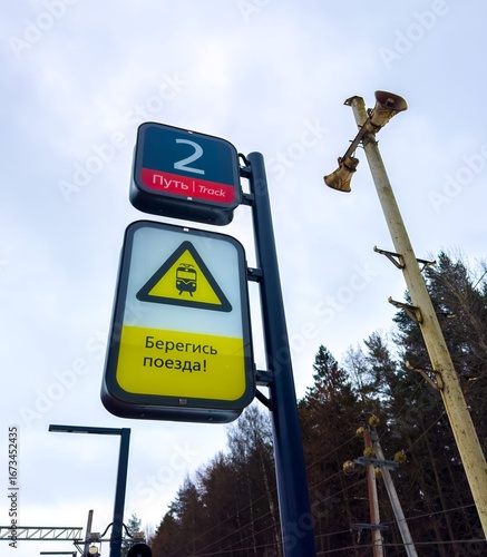 A low-angle shot of two signs on a platform. The top sign indicates Track 2 in Russian and English. Below it, a bright yellow triangular warning sign with a train icon warns Beware of the train in