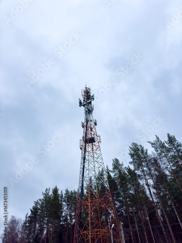 A dramatic, low-angle shot of a tall telecommunication tower with red and white sections, extending high into a cloudy sky. The steel lattice structure is framed by the tops of pine trees on either