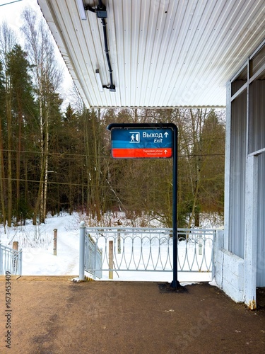 A medium-shot captures a directional sign with a person icon and arrow pointing to the Exit (Vykhod) to a street. The sign stands on a snow-covered train platform, with a white, ribbed roof overhead
