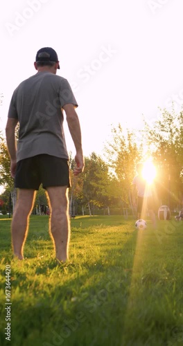 Happy father teaching son how to pass football in park at sunset. Dad shows boy how to kick soccer ball while practicing. Father and child play sports outdoors, strengthening family bond. Vertical