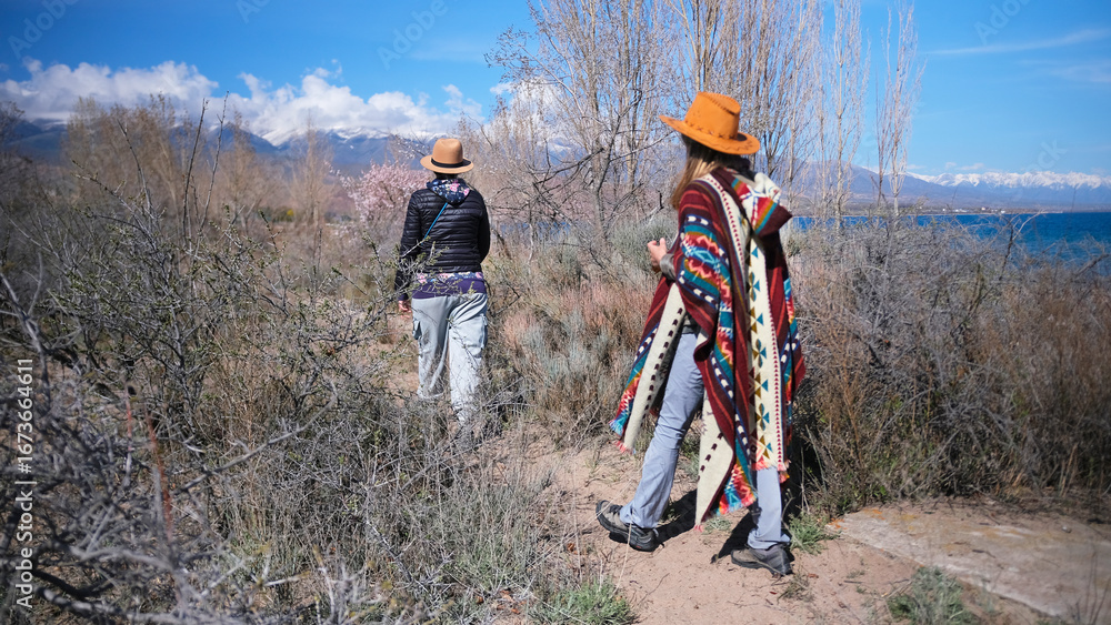 Obraz premium Two women are walking through a field overgrown with dry bushes. Two friends are walking near the blue lake. Travel and adventures