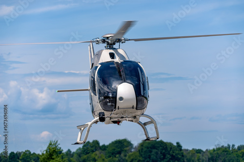 A white Airbus H120 helicopter hovers front-on above the runway, skids down and rotor blades spinning, framed by green treetops and a layered sky filled with altostratus and cumulus clouds.