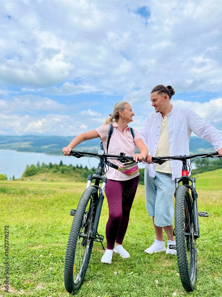 Obraz premium Happy smiling couple with bicycles on a cycling path near Czorsztyn and Czorsztynskie Lake with Tatra Mountains in the background, Poland. Concept of active lifestyle, love, adventure, and eco tourism