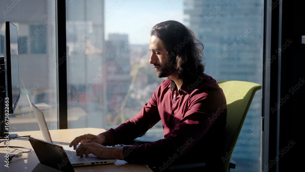 Fototapeta premium Millennial caucasian man working on a laptop by a large window in a bright modern office, focused and productive, highlighting technology, communication and professional dedication indoors.