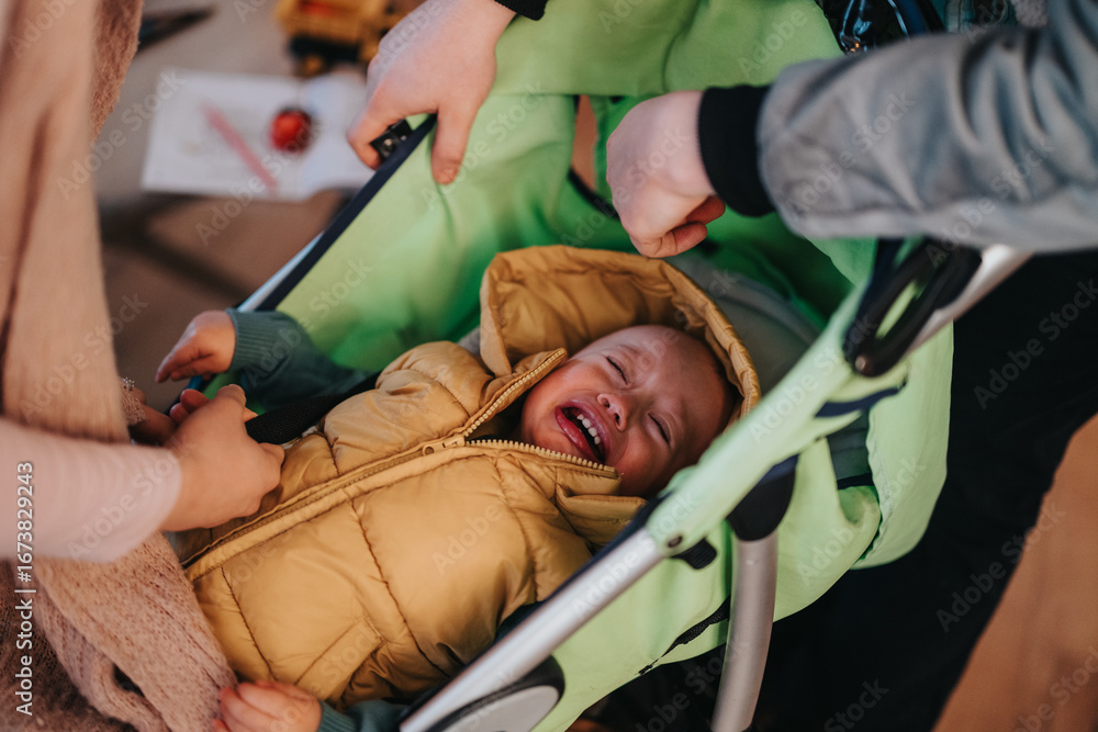 Obraz premium A scene depicting a young child crying while two adult hands prepare them in a stroller. The situation portrays childhood emotions and parental care.