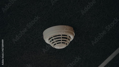 A white smoke detector is attached to a textured black ceiling. The detector is a circular shape with a domed cover and vented sides. It represents safety, protection, fire prevention in any home