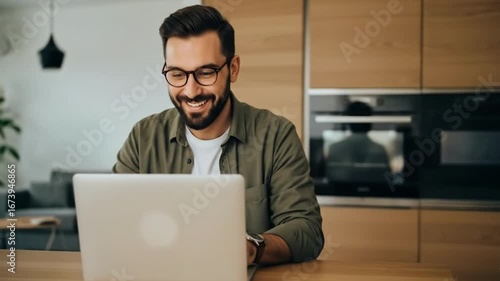Smiling man with glasses working on a laptop at a table in a modern kitchen