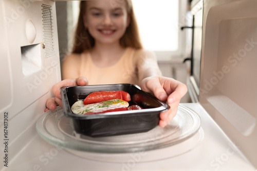 Smiling girl putting ready meal tray into microwave oven, view from inside the appliance.