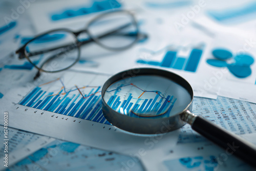 Magnifying glass over financial charts and graphs with eyeglasses in background for business analysis