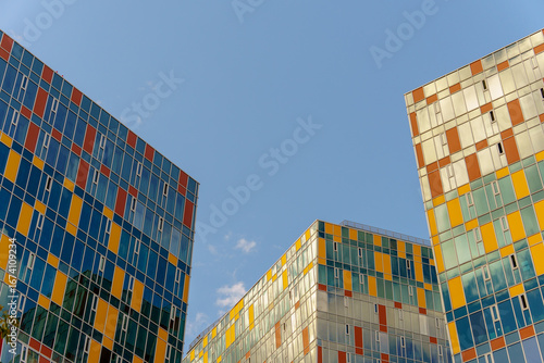 Three high-rise buildings with colorful panel facades set against a clear blue sky. Bright tones and reflective surfaces create a dynamic urban composition.