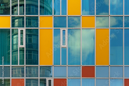 Facade of a modern building featuring a grid of rectangular panels in blue, yellow, and reddish-orange. Glass sections reflect the sky and clouds, adding a dynamic visual effect.