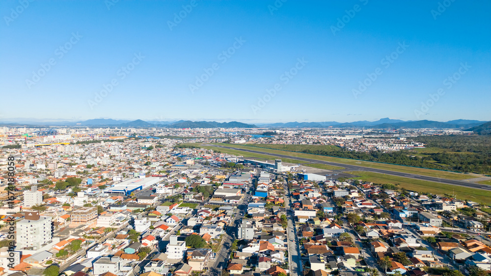 Fototapeta premium Aerial view of Navegantes Airport runway surrounded by city buildings on a sunny day.