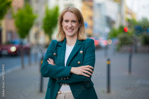 Confident businesswoman in green blazer smiling with arms crossed outdoors in city street environment  
