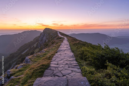 A narrow, winding stone path leads along a majestic mountain ridge, illuminated by the warm glow of a sunset. 