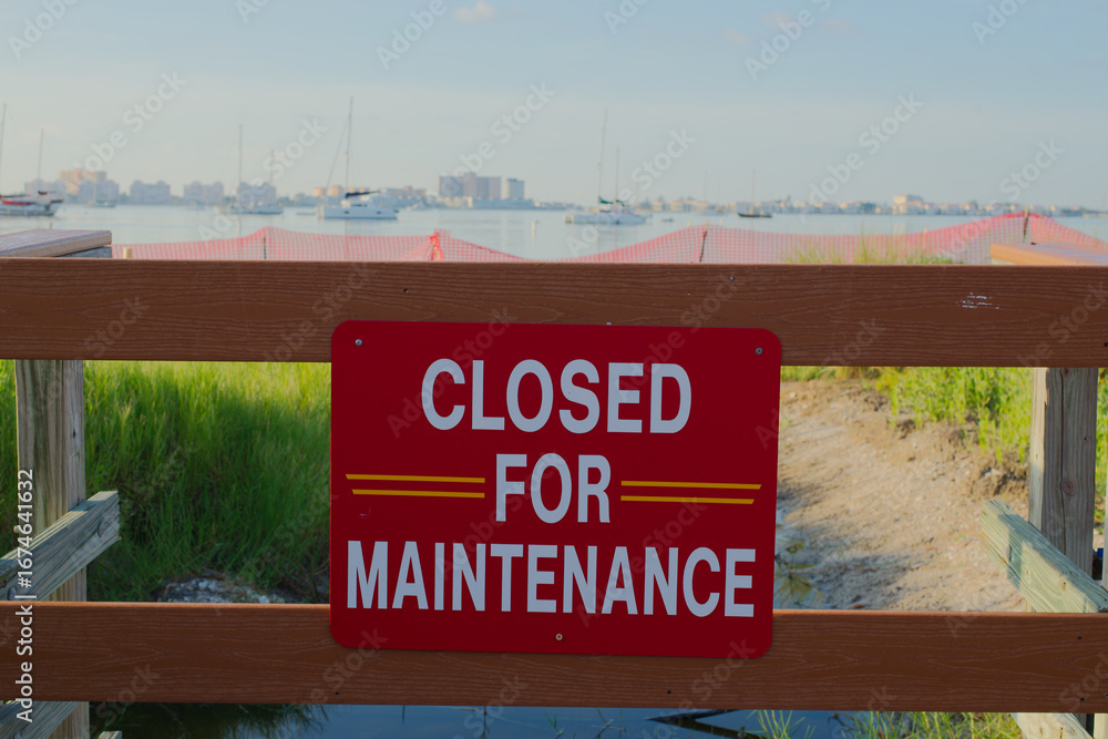 Obraz premium Maintenance Closure Sign Overlooking Coastal Scenery and Boats Gulfport, FL. Wooden fencing, with a scenic background of grassy coastline, water, and distant boats under a clear sky. 