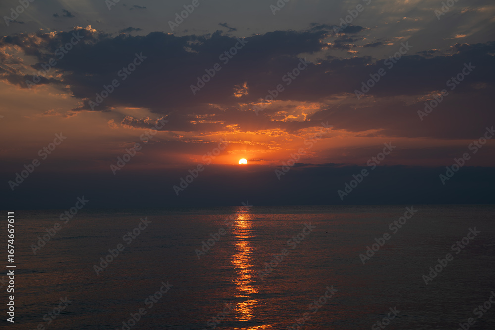 Obraz premium Dramatic golden sunrise over sea with breaking through dark clouds. Peaceful and serene horizon at dawn as the sun rises and reflects on calm water. Hope and new beginnings