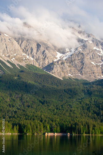 Eibsee Lake in Bavaria, Germany, with small lakeside houses and cabins at the forest edge, beneath the towering Zugspitze mountains, partly covered by clouds. A scenic alpine landscape.