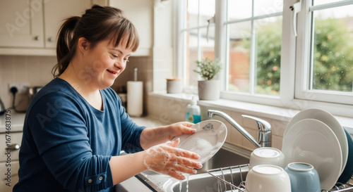 Happy woman with down syndrome washing dish in kitchen sink. Daily routine and independent living concept for disability awareness.
