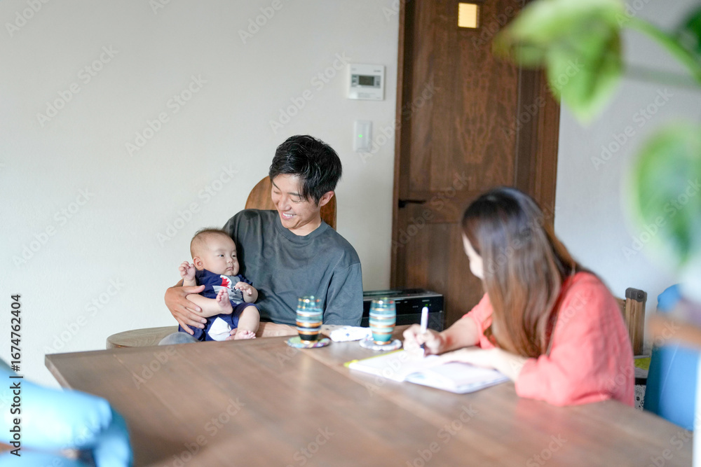 Fototapeta premium A man and a woman are sitting at a table with a baby in front of them. The man is holding the baby while the woman is writing something. Scene is warm and loving