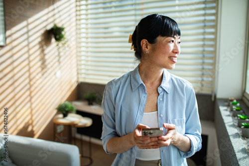 Asian woman using smartphone at home while standing by window blinds