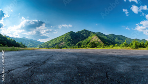 Slika na platnu An open asphalt area with a beautiful mountainous landscape under a clear blue sky with scattered clouds