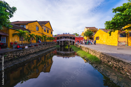 A scenic view of the iconic Chua Cau bridge reflecting on a small canal, surrounded by traditional yellow buildings and lush greenery on a peaceful day in Hoi An.