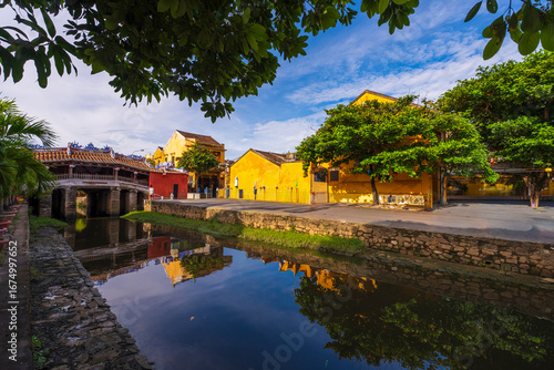 A scenic view of the iconic Chua Cau bridge reflecting on a small canal, surrounded by traditional yellow buildings and lush greenery on a peaceful day in Hoi An.