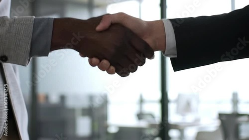 Two people one with dark skin and wearing a gray jacket and another wearing a black suit shaking hands in a bright office space