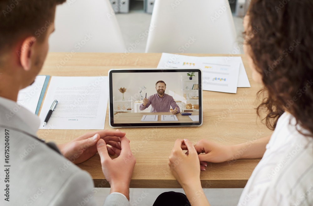 Fototapeta premium Two young business people sitting at the desk in office having online video call with male colleague using tablet. Coworkers and company employees chatting online during video conference.