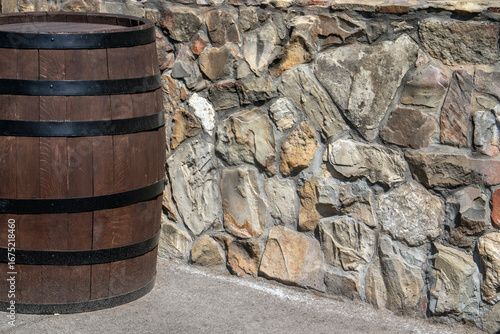 Empty oak barrel stands against a stone wall, illuminated by the sun. Wine barrel with a black steel hoop. Metal and natural wood of a wine barrel, view on a sunny day.