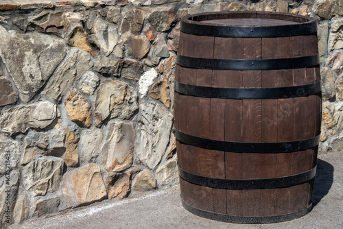 Empty oak barrel stands against a stone wall, illuminated by the sun. Wine barrel with a black steel hoop. Metal and natural wood of a wine barrel, view on a sunny day.