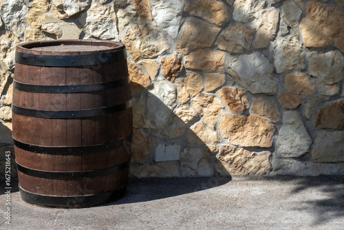 Empty oak barrel stands against a stone wall, illuminated by the sun. Wine barrel with a black steel hoop. Metal and natural wood of a wine barrel, view on a sunny day.