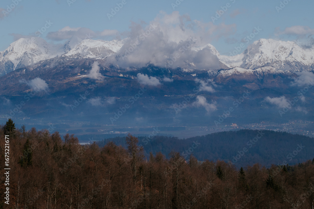 Obraz premium Majestic Kamnik Alps rise above a misty valley near Ljubljana, Slovenia, with rugged peaks and snow-capped summits glowing under the early morning light
