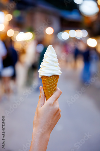 A close-up of a hand holding a soft serve ice cream cone against a blurry background of a charming street in Mae Kampong, Thailand, representing a local travel experience.
