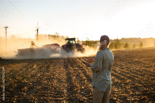 Farmer with smartphone monitoring smart farming cultivation while tractor and seeder prepare soil during agricultural work on field