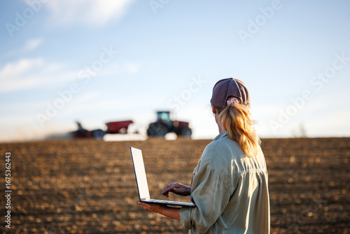 Smart farming. Woman farmer with laptop analyzing agricultural data in front of tractor and seeding machine on cultivated field