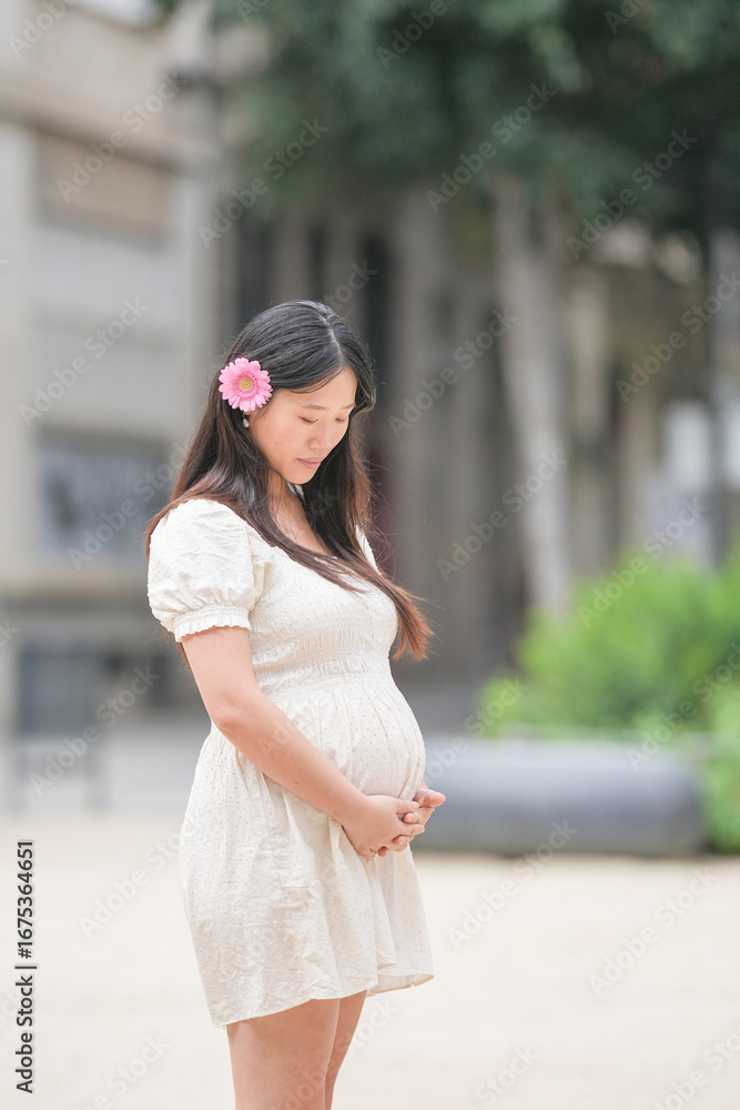 Fototapeta premium A woman is standing in a white dress with a pink flower in her hair. She is pregnant and looking at the camera