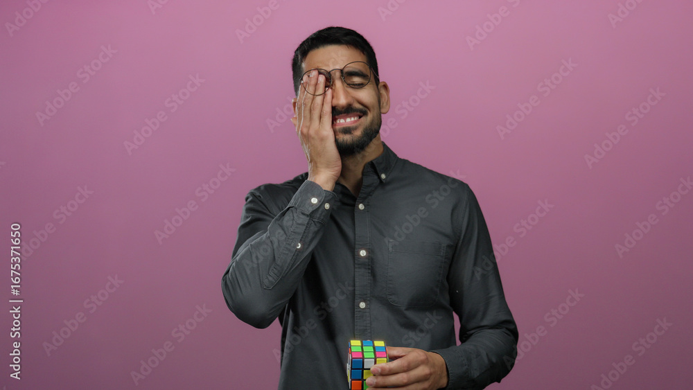 Φωτογραφία Young man holding cube smiling over pink background, wearing glasses and black shirt, expressing happiness and contemplation