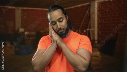 Obraz na plátně Man wearing bright orange shirt with hands pressed to cheek inside building under construction, eyes closed and head tilted; work fatigue