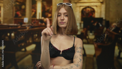 Obraz na plátně Young blonde tattooed woman wearing black bikini pointing finger in church interior pews; rebuke defiance