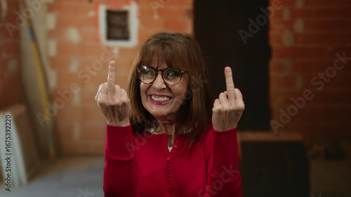 Billede på lærred Senior hispanic woman showing middle fingers indoors at a construction site with a mischievous expression against a brick wall background