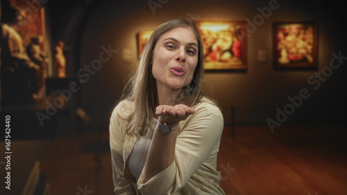 Young blonde woman softly blows kiss with hand and smiles at camera under warm gallery lights in building; affection.