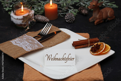 Tischdekoration zur Weihnachtsfeier. Kerzen mit Tannenzweigen und ein Tischgedeck. Auf einer Karte steht der Text Weihnachtsfeier.