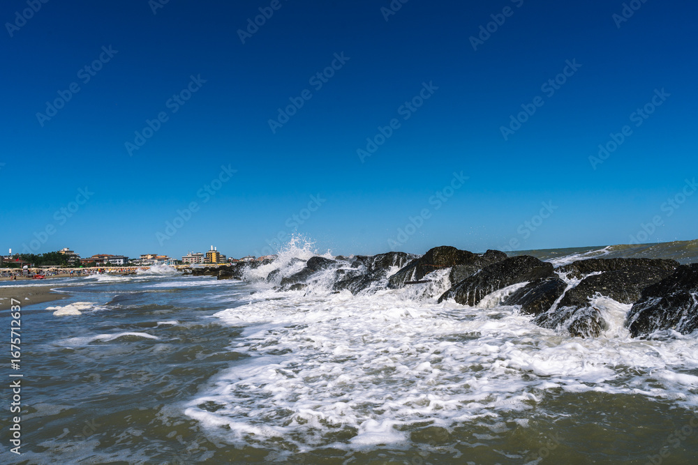 Fototapeta premium Waves crashing against rocks near a beach, creating a dynamic seascape against a clear blue sky. Coastal beauty unfolds.